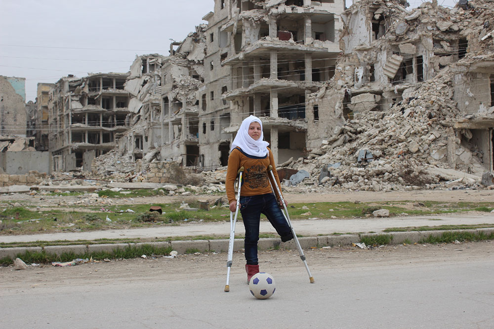 En jente med ett ben spiller fotball med krykker foran ruiner i Syria