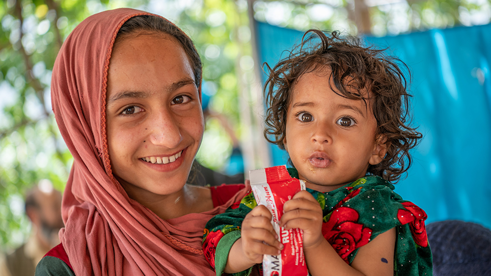 Et barn fra Afghanistan spiser fra nødmatpakker. Et eldre barn står ved siden av og smiler. FOTO