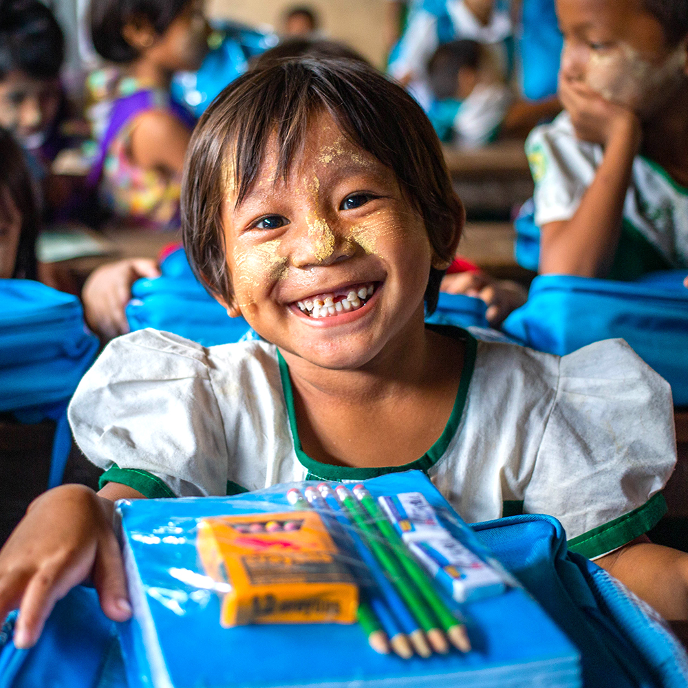 Ei jente smiler mot kamera med skolesaker foran seg, Myanmar