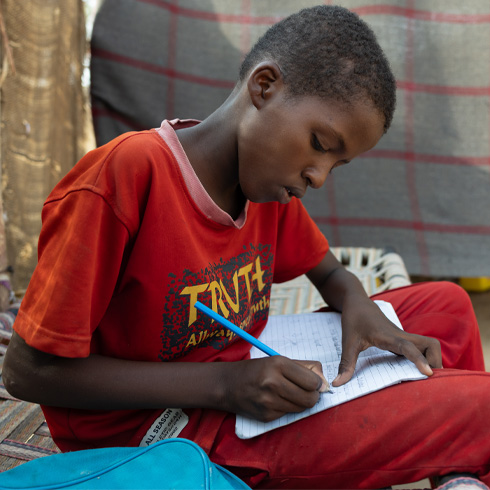 En gutt sitter på en feltseng og gjør hjemmelekser, Sudan