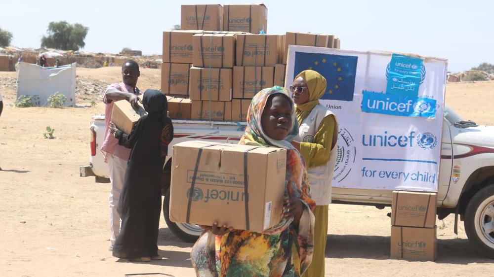 En kvinne bærer en UNICEF-eske, bak henne står en bil med mange esker, Sudan