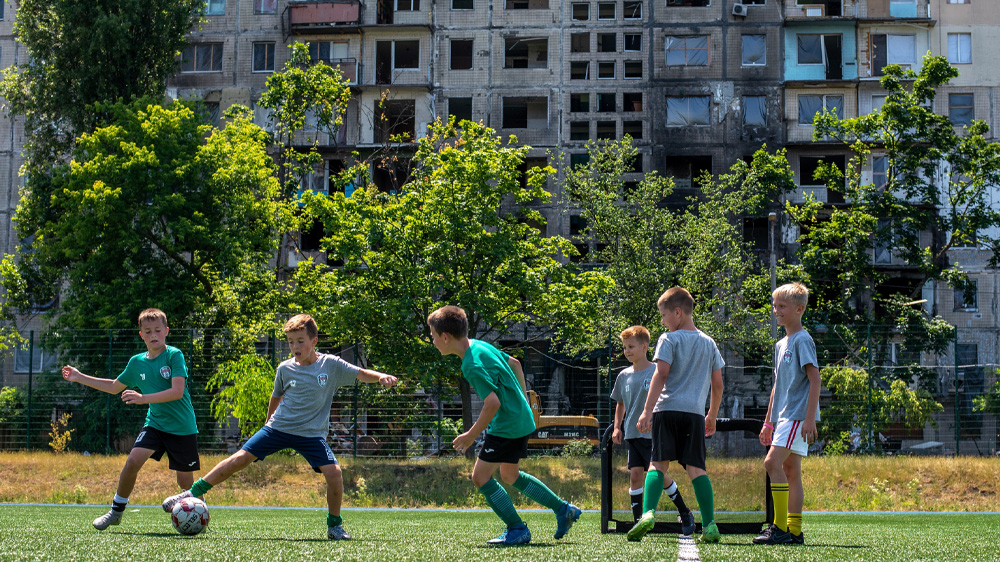 En gruppe barn spiller fotball foran bombede bygninger, Ukraina