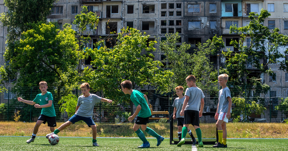 En gruppe barn spiller fotball foran bombede bygninger, Ukraina