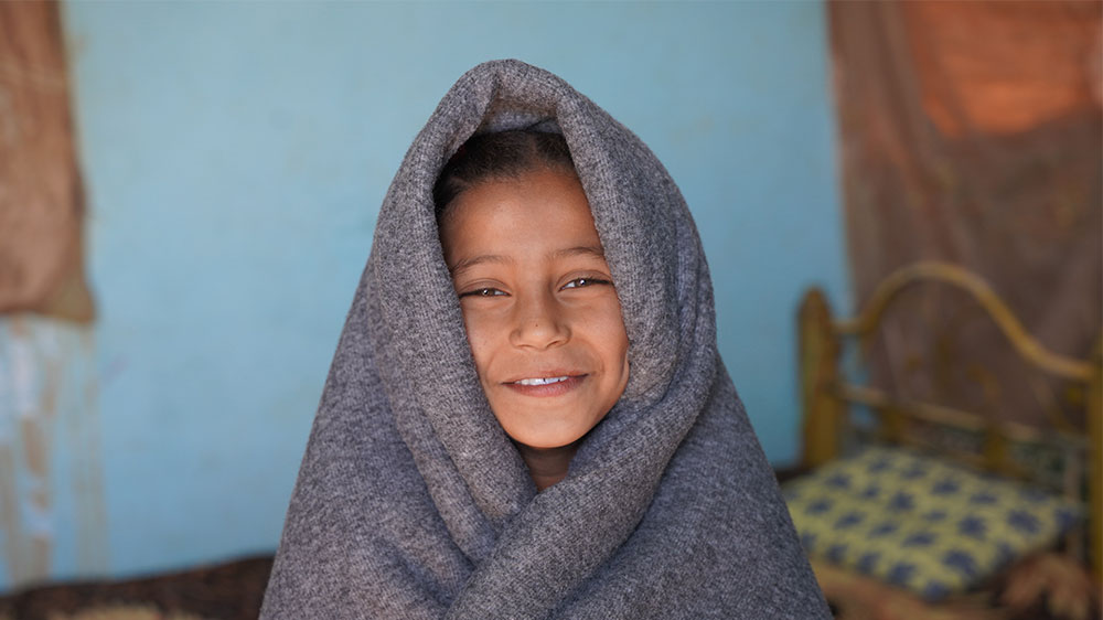 En jente i Sudan smiler og har et varmt pledd rundt seg.