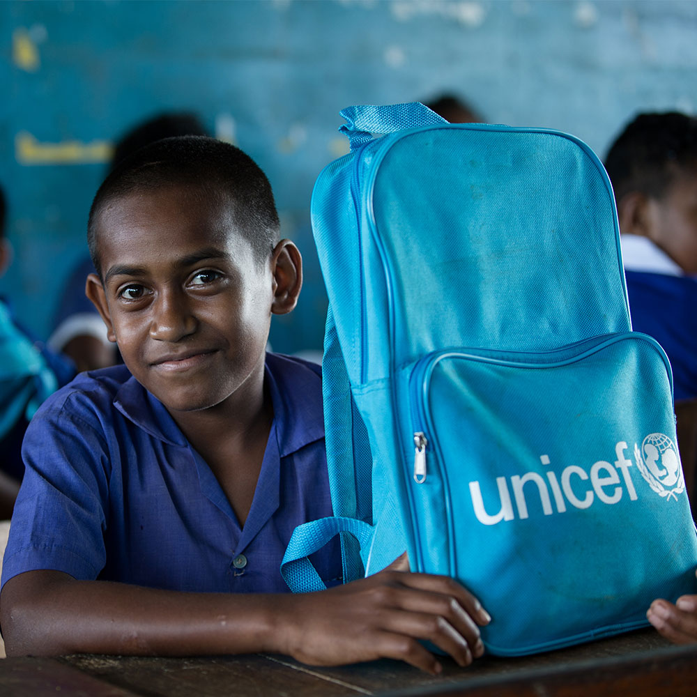 En gutt smiler i kamera med en sekk fra UNICEF på pulten