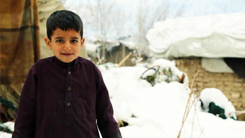 Afghansk, tynnkledd gutt ser inn i kamera. Rundt ham ser vi falleferdige hus og snø.