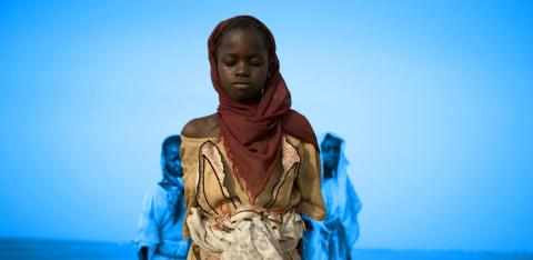 En ung jente iført hodeplagg og en slitt kjole står og ser ned mot bakken, to andre jenter går bak henne. 