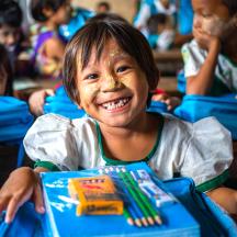 Ei jente smiler mot kamera med skolesaker foran seg, Myanmar